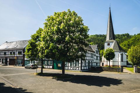 Ein malerisches Dorf mit traditionellen Fachwerkhäusern und einem hohen Kirchturm. Im Vordergrund steht ein großer, blühender Baum.
