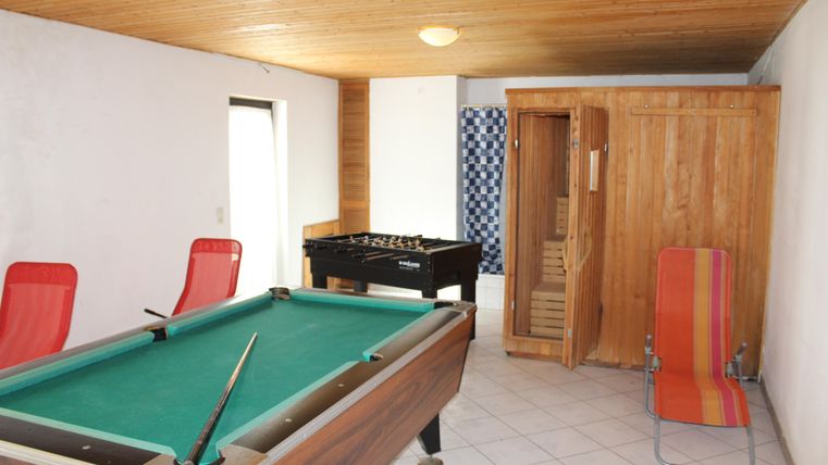 Ein gemütlicher Freizeitraum mit einem Billardtisch, einem Tischfußballspiel und einem Sauna-Bereich. Zwei rot-orange Stühle laden zum Entspannen ein.