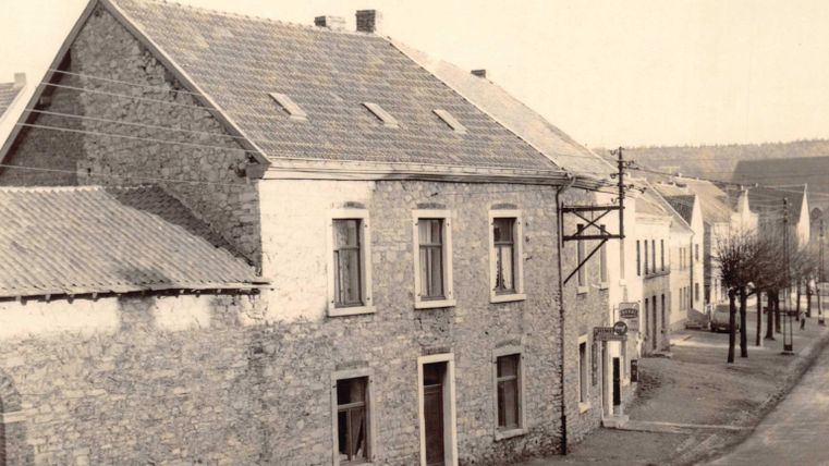 Ein altes, steinernes Haus an einer ruhigen Straße. Im Hintergrund sind mehrere weitere Gebäude und Bäume sichtbar.