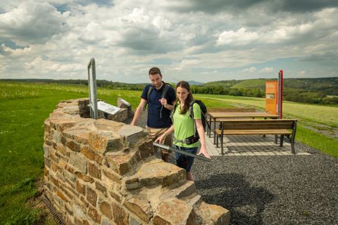 A viewpoint with two people standing by a rock formation. In the background, green hills and a cloudy sky can be seen.