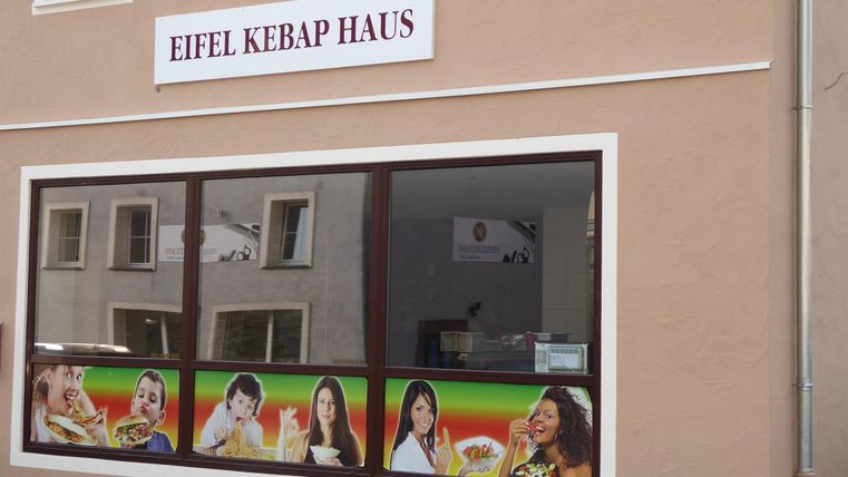 Ein Restaurant namens "Eifel Kebap Haus" mit großen Fenstern. Die Fassade zeigt Bilder von verschiedenen Gerichten und fröhlichen Menschen.