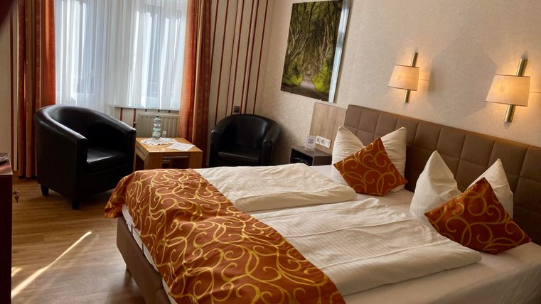 Ein gemütliches Hotelzimmer mit zwei Betten und orangefarbenen Dekokissen. Im Hintergrund sind zwei Sessel und ein Tisch sichtbar.