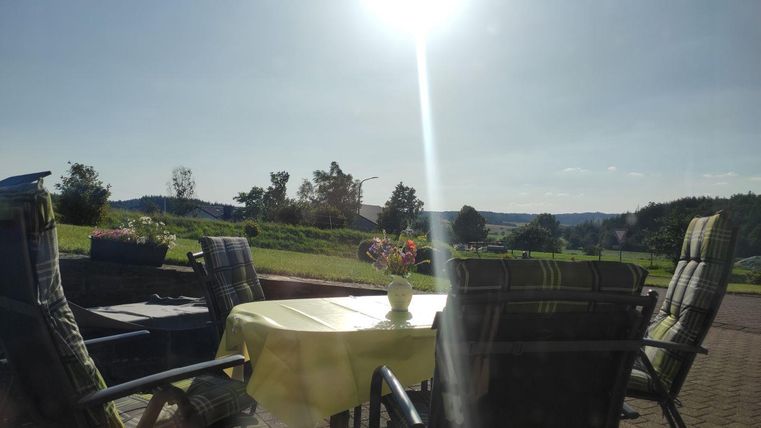 Ein sonniger Außenbereich mit einem Tisch und mehreren Stühlen. Im Hintergrund sind grüne Wiesen und Bäume zu sehen.