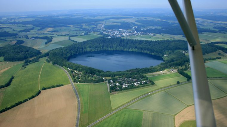 Een landschap met een ronde meer, omringd door groene velden en bossen. Deels beboste heuvels en een klein dorp zijn ook zichtbaar.