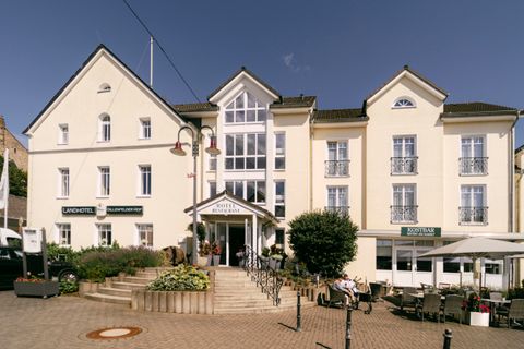 Ein einladendes Hotel mit einer modernen Fassade. Vor dem Gebäude sind Tische im Freien und Blumenbeete zu sehen.
