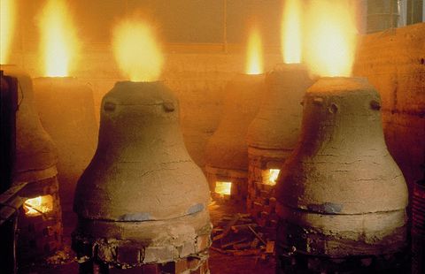 In einem alten Ofenraum stehen mehrere große Öfen, aus denen Flammen aufsteigen. Die Wände sind aus Stein und die Atmosphäre ist warm und rauchig.