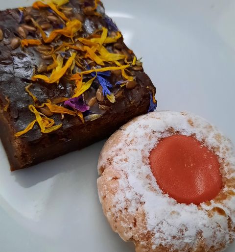 Ein Stück Schokoladenkuchen mit bunten Blütenblättern und ein Keks mit einer roten Marmeladefüllung. Beide Leckereien sind auf einem weißen Teller angerichtet.