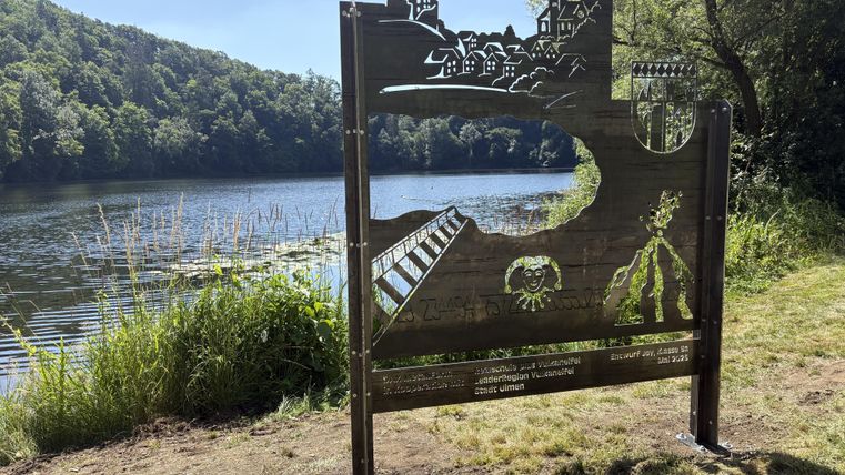 Ein kunstvolles Blickfang mit einer Darstellung von Landschaft und Architektur am Wasser. Im Hintergrund sind Bäume und ein ruhiger See zu sehen.