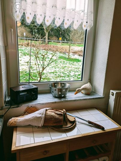 A cozy kitchen nook with a window that looks out onto a winter garden. On the table are a bread knife, a loaf of bread, and a tablecloth.