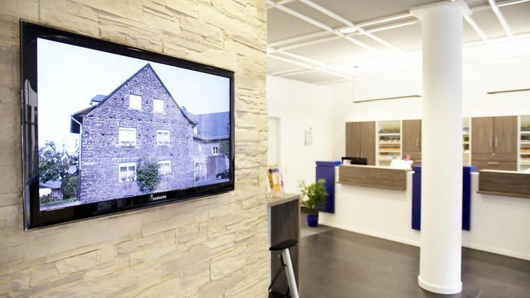 Een moderne wachtkamer met een receptiebalie en een grote muur met een televisie. De televisie toont een afbeelding van een gebouw.