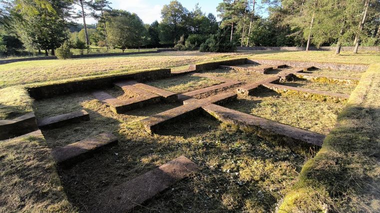 Ruinen eines alten Gebäudes auf einer Wiese, umgeben von Bäumen. Die Struktur ist rechteckig und aus Stein.