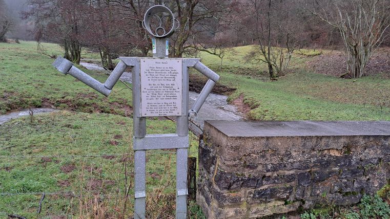 A statue made of metal in the shape of a person stands in a green landscape. In the background, a small stream flows and some trees are visible.