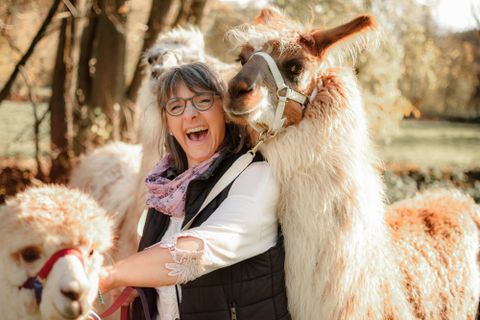Eine fröhliche Frau umarmt ein Lama und lächelt in die Kamera. Im Hintergrund sind weitere Lamas und herbstliche Bäume zu sehen.