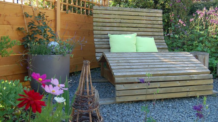 A cozy wooden lounge chair is located in a blooming garden. Colorful flowers and a beautiful garden structure surround the relaxation area.