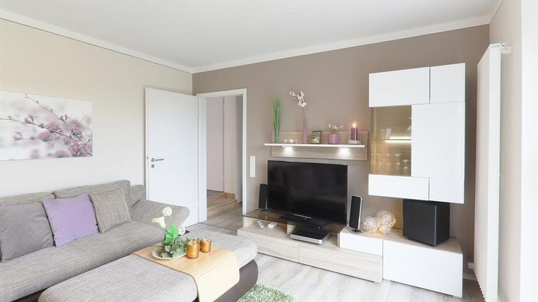 Een moderne woonkamer met een grijze bank en een televisie op een helder TV-meubel. De muren zijn in neutrale kleuren gehouden, en er zijn decoratieve elementen zoals planten en een tapijt.