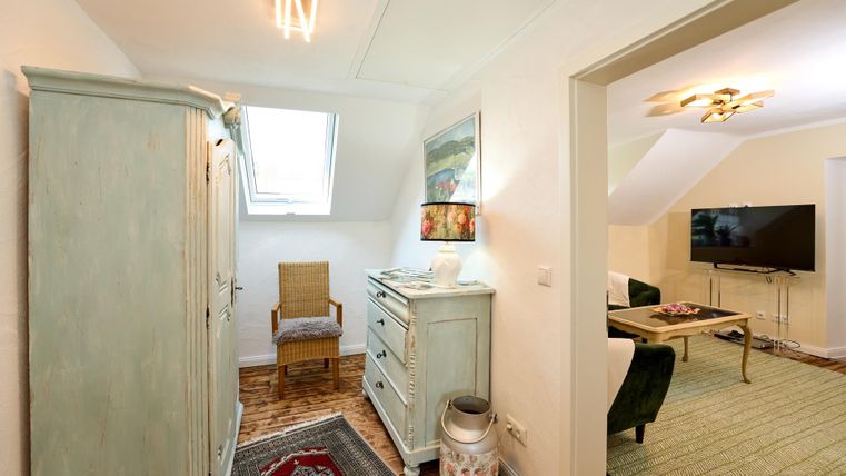 A cozy hallway with a light green cupboard and a chair. In the background, a living area with a sofa and TV is visible.