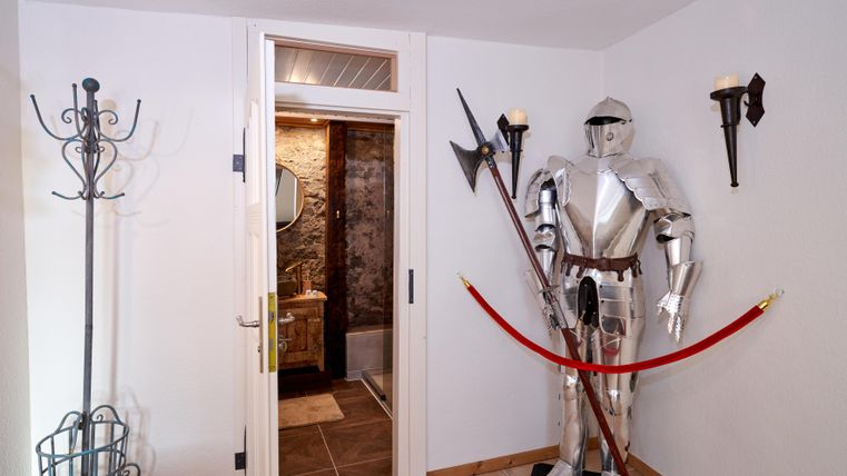 A knight in full armor stands by a wall next to a door. The room has bright walls and some wall lamps.
