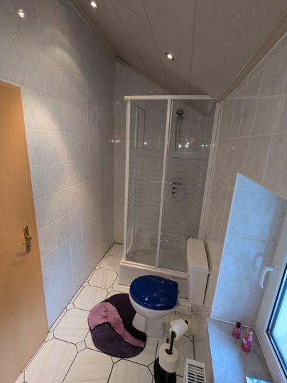 A modern bathroom with a shower and a blue toilet. The walls are light tiled and a round rug is on the floor.