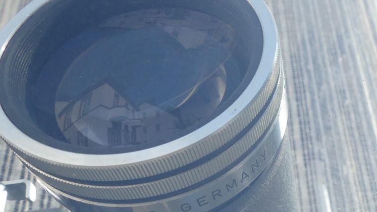 Ein Kameraobjektiv mit einem klaren Glanz und der Aufschrift "Köln, Deutschland". Im Hintergrund sind reflektierte Gebäude zu sehen.