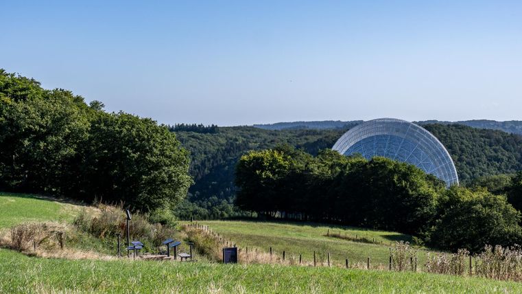 Eine grüne Wiese mit Bäumen im Vordergrund und einem großen geodätischen Dome im Hintergrund. Der Himmel ist klar und blau.