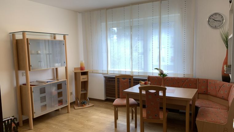 Ein helles Wohnzimmer mit einem Essbereich und einem großen Fenster. Es gibt einen Tisch, Stühle und ein Regal mit Glasfront.