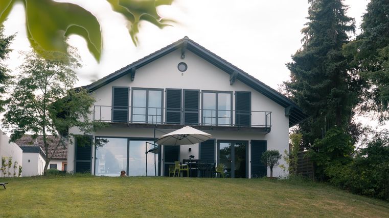 Ein modernes Haus mit einer großen Terrasse und einem Gartentisch. Der Garten ist grün und von Bäumen umgeben.