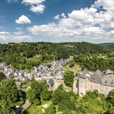Panoramablick auf Monschau mit der Burg im Vordergrund, umgeben von grüner Landschaft und bewaldeten Hügeln unter blauem Himmel mit Wolken., © Eifel Tourismus GmbH, Dominik Ketz