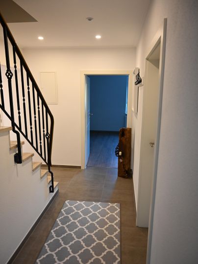 Een lichte gang met een trap en een decoratief tapijt. Op de achtergrond is een open deur die naar een andere kamer leidt.