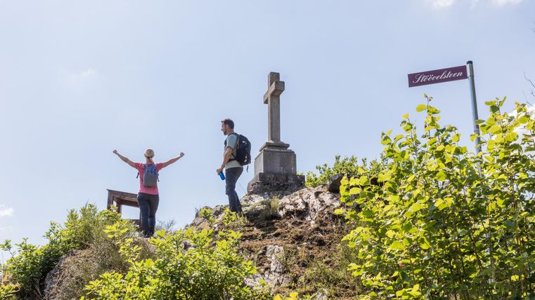A woman stands with her arms outstretched on a hill, while a man stands next to her. In the background, a cross and a signpost can be seen.