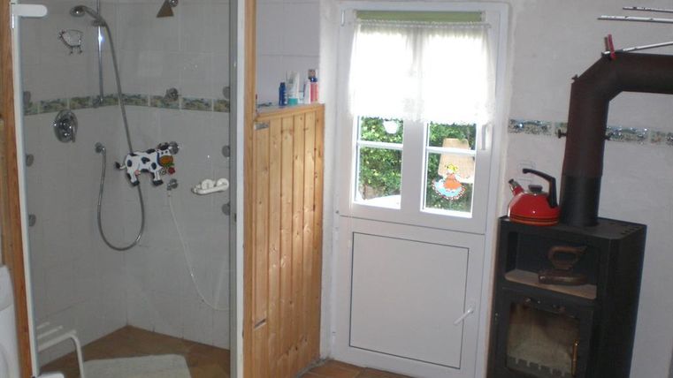 Ein Badezimmer mit einer Dusche, die durch eine Glaswand abgetrennt ist. Neben der Dusche steht ein Kaminofen, und es gibt ein Fenster mit Vorhängen.