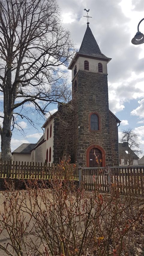 Eine kleine Kirche mit einem spitzen Turm und einem schönen roten Eingang. Im Vordergrund sind trockene Büsche und ein Zaun sichtbar.