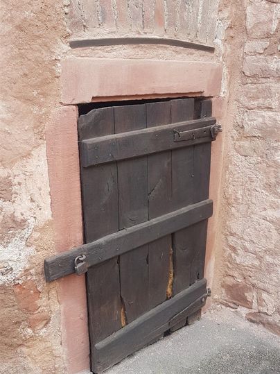 Eine kleine Holztür in einer Steinmauer. Die Tür ist dunkelbraun und hat eine rustikale, antike Optik.