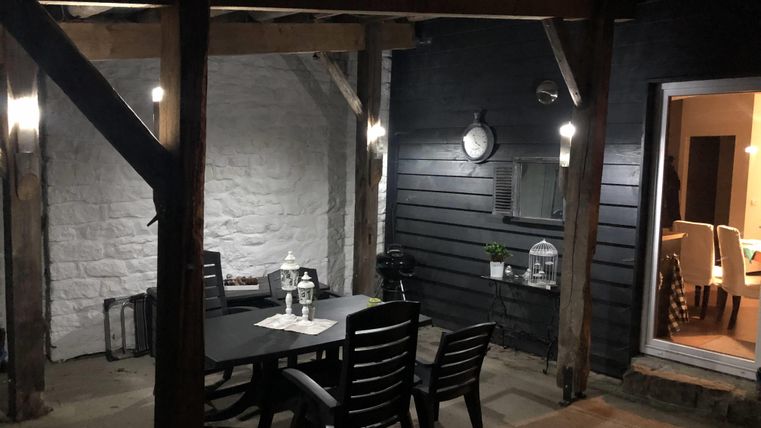 Ein gemütlicher Außenbereich mit einem Tisch und Stühlen unter einem Holzüberhang. Die Umgebung ist schwach beleuchtet und bietet Zugang zu einem Innenraum.