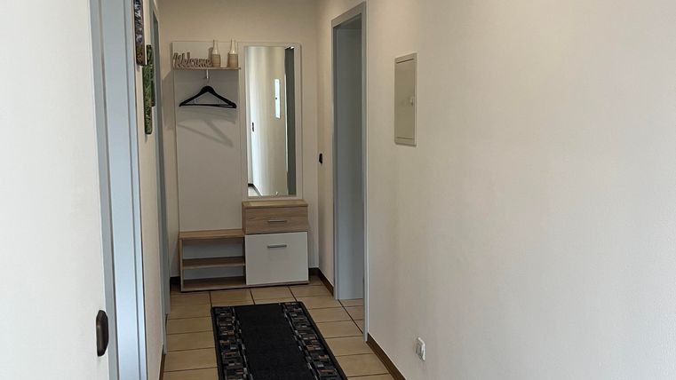 A bright hallway with white walls and a long carpet. At the end of the hallway, there are cabinets and a coat hook.
