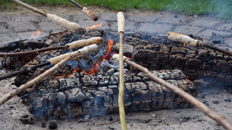 Een kampvuur met verbrande houtblokken staat in het middelpunt. Stokken met deeg worden boven de vlammen gehouden.