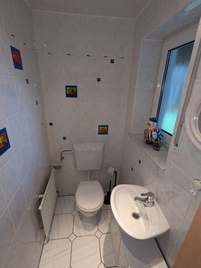 A small, modern bathroom with white tiles. It includes a sink, a toilet, and a window.