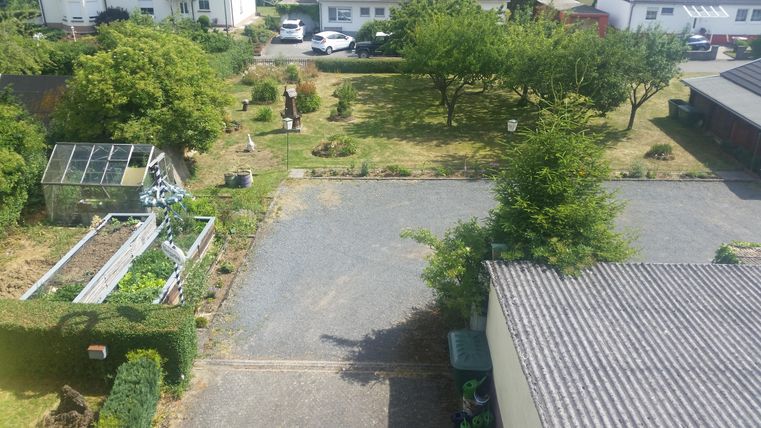 Eine ruhige Auffahrt mit grauem Kies und einem Gartenbereich. Im Hintergrund sind Bäume und einige Autos zu sehen.