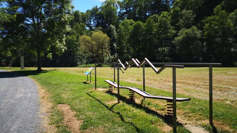 Een fitnessruimte in de buitenlucht met turntoestellen op een groene weide. Op de achtergrond zijn bomen en een blauwe lucht te zien.