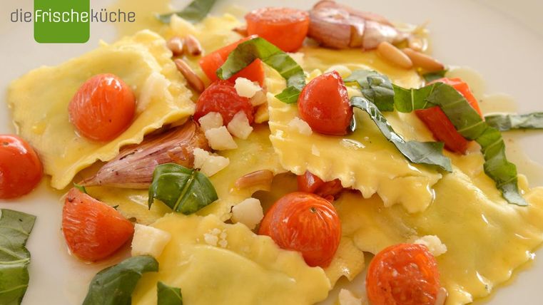 Ein Teller mit frischen Ravioli, angereichert mit Kirschtomaten, Basilikum und Pinienkernen. Die Farben sind lebhaft und appetitlich.
