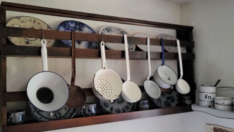 A wooden shelf displays various kitchen utensils, including strainers and bowls. In the background, decorative plates and pots can be seen.