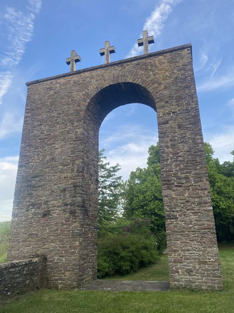 Blick auf einen  gemauerten Torbau etwas 10m hoch.Die rechte Wand ist breiter als die linke Wand. Auf dem geraden Abschluss stehen 3 Kreuze. Der Torbau steht auf einer Freifläche. Hinter dem Torbau sieht man einige Bäume.