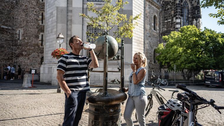 Ein Mann trinkt Wasser an einem Brunnen, während eine Frau lächelnd daneben steht. Im Hintergrund sind Fahrräder und eine historische Architektur zu sehen.