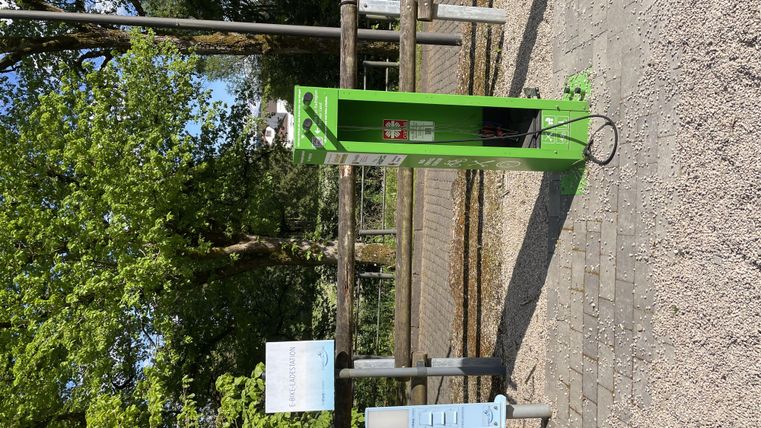 Eine grüne Ladestation für E-Bikes steht auf einem Weg umgeben von Bäumen. Neben der Station befindet sich ein Informationsschild.