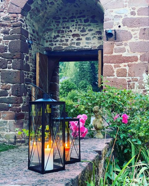 A beautiful lantern stands in front of an old stone archway. In the background, green plants and colorful flowers can be seen.
