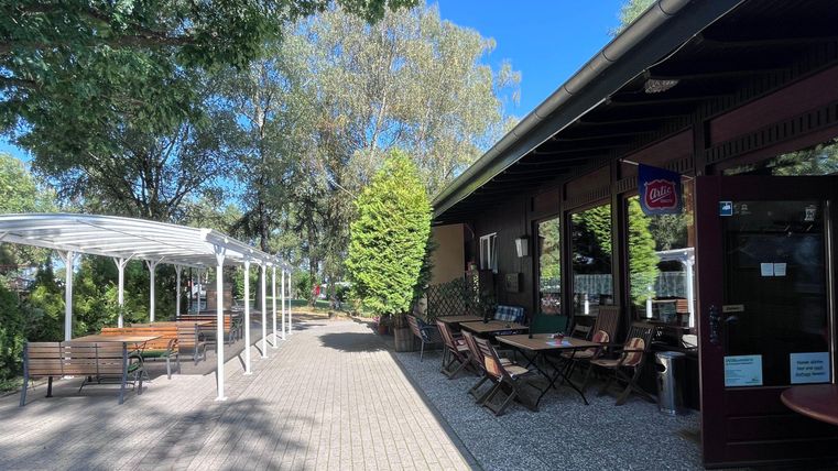 Een gezellige buitenruimte met tafels en banken onder bomen. Op de achtergrond is een gebouw met ramen en een ingang te zien.