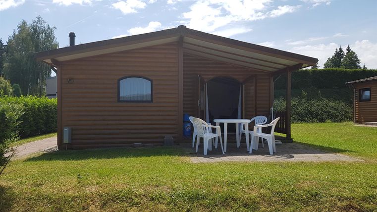 Ein Holzbungalow mit überdachter Terrasse, weißen Plastikstühlen und Tisch auf einer grünen Wiese.