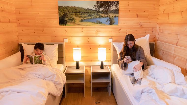 Ein gemütliches Zimmer mit Holzverkleidung und zwei Einzelbetten. Ein Kind liest ein Buch, während ein Jugendlicher auf dem Handy sitzt.