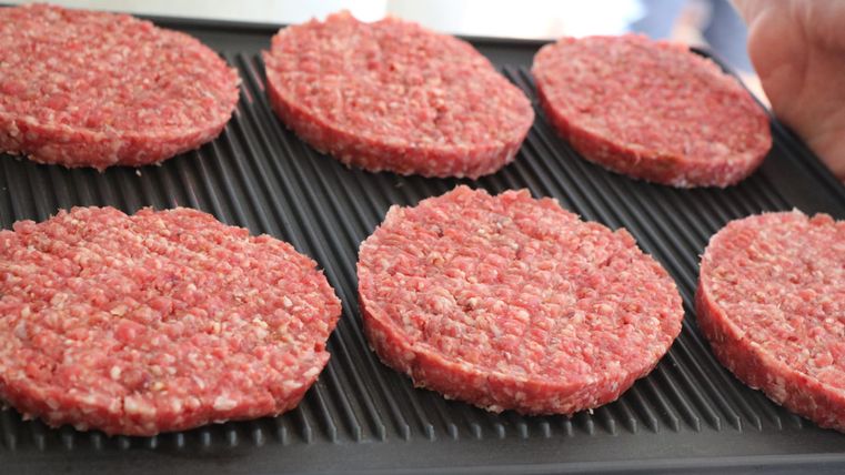 Sechs rohe Hackfleisch-Patties liegen auf einem Grill, bereit zum Braten. Die Patties sind gleichmäßig geformt und von roter Farbe.