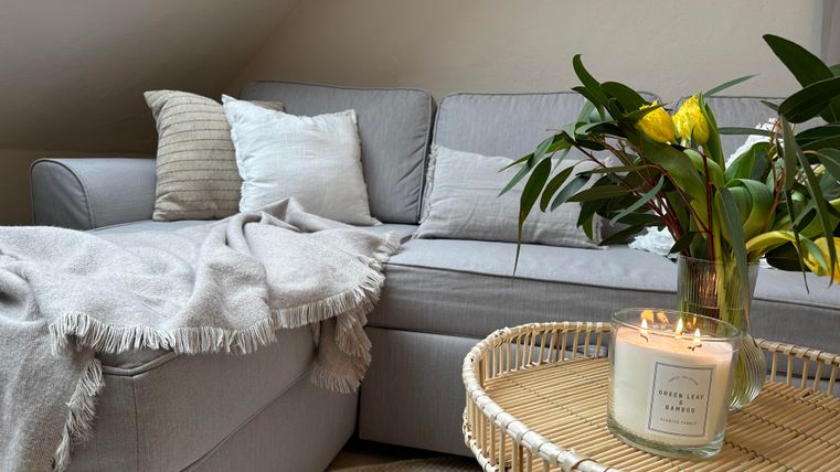 Een gezellige woonkamer met een grijze bank en kussens. Op de tafel staat een vaas met verse bloemen en een kaars.