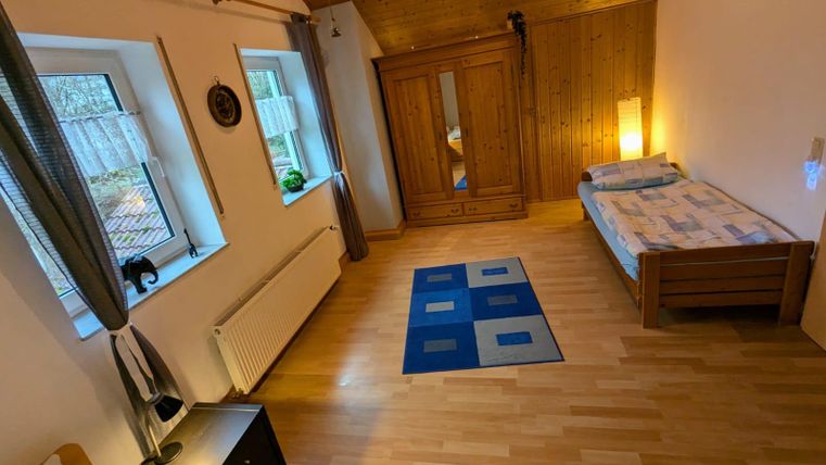 A cozy room with two single beds and a wooden wardrobe. Large windows let in plenty of daylight and the floor is covered with wood.